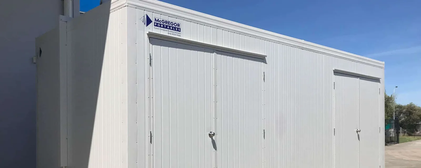 White portable building viewed from the corner, with the McGregor Portables sign and phone number near the roofline, under blue sky