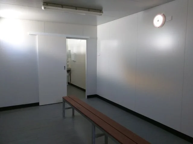 Interior of a changeroom, with white walls, a timber bench seat running along one wall and an open doorway through to a further room.