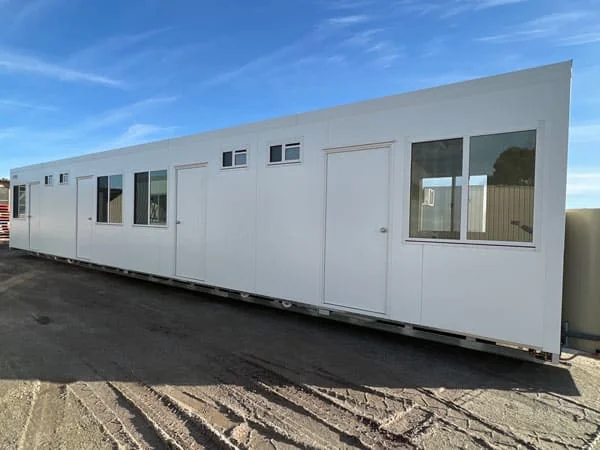 Long white bunkhouse on a dirt yard, with a run of doors and windows along one side.