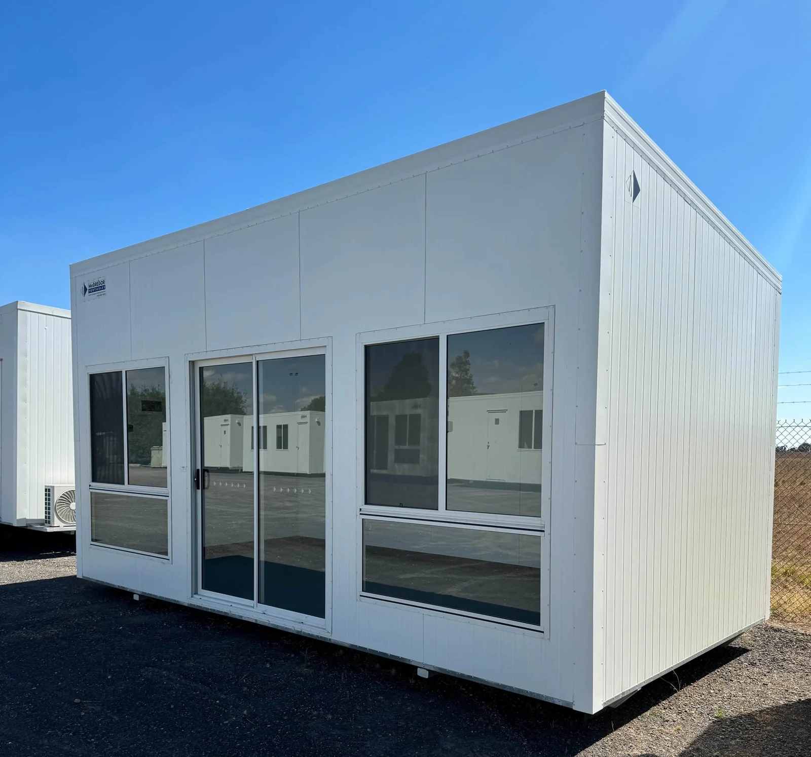 White sales office with sliding glass entry doors set between two large windows, on a gravel yard.