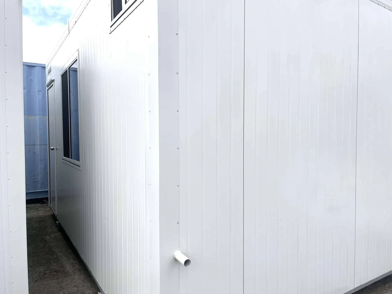 Side wall of a used white site office, largely blank cladding with a short pipe outlet near the ground and another building behind.