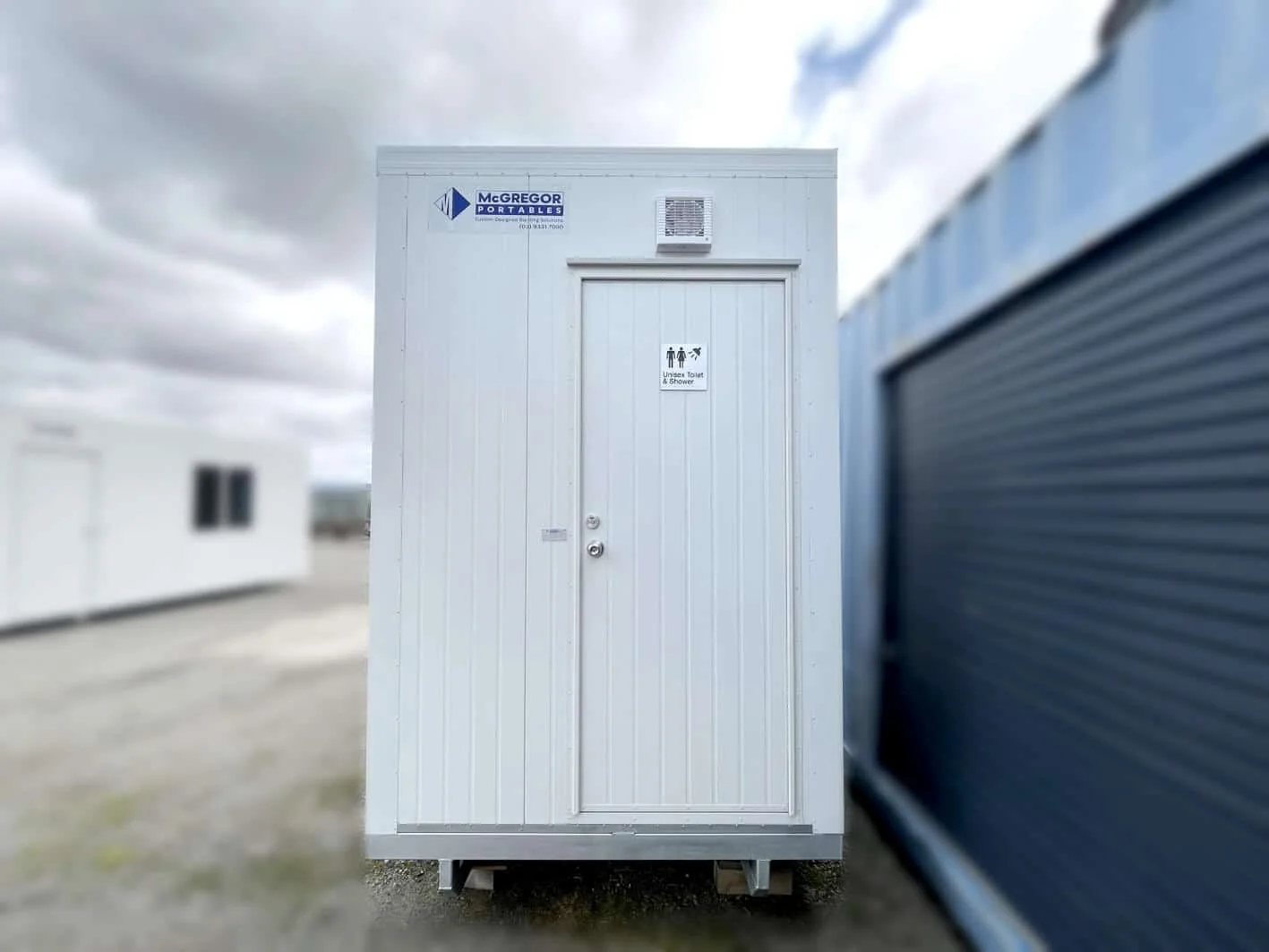 Small white ensuite unit with a single signed door, standing beside a corrugated shed.