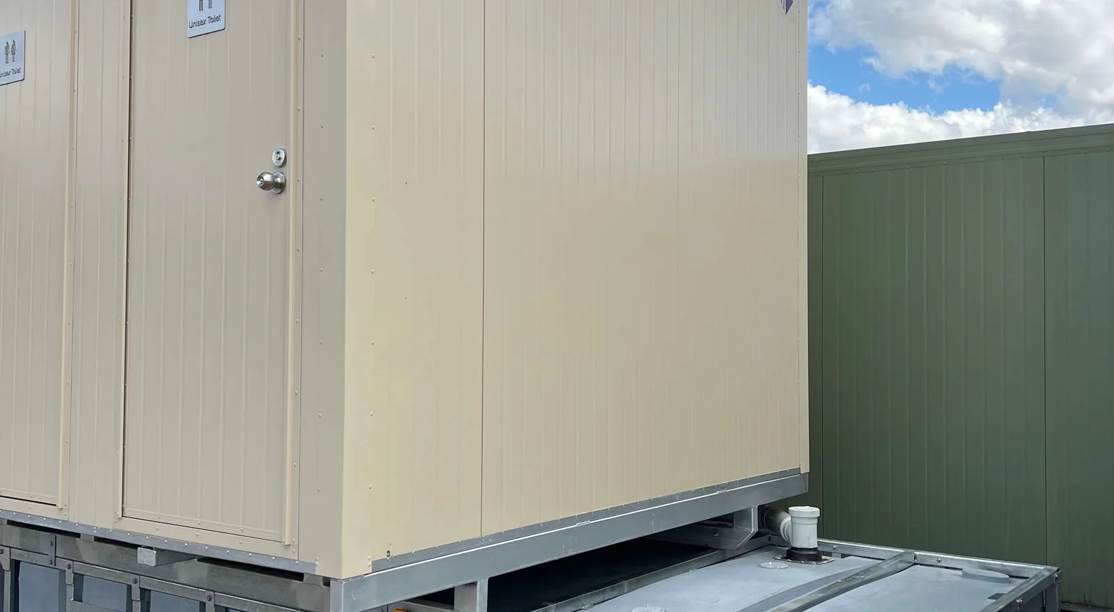 Cream unisex toilet block with two signed doors, raised on a galvanised steel frame with the waste storage tank sitting alongside underneath.