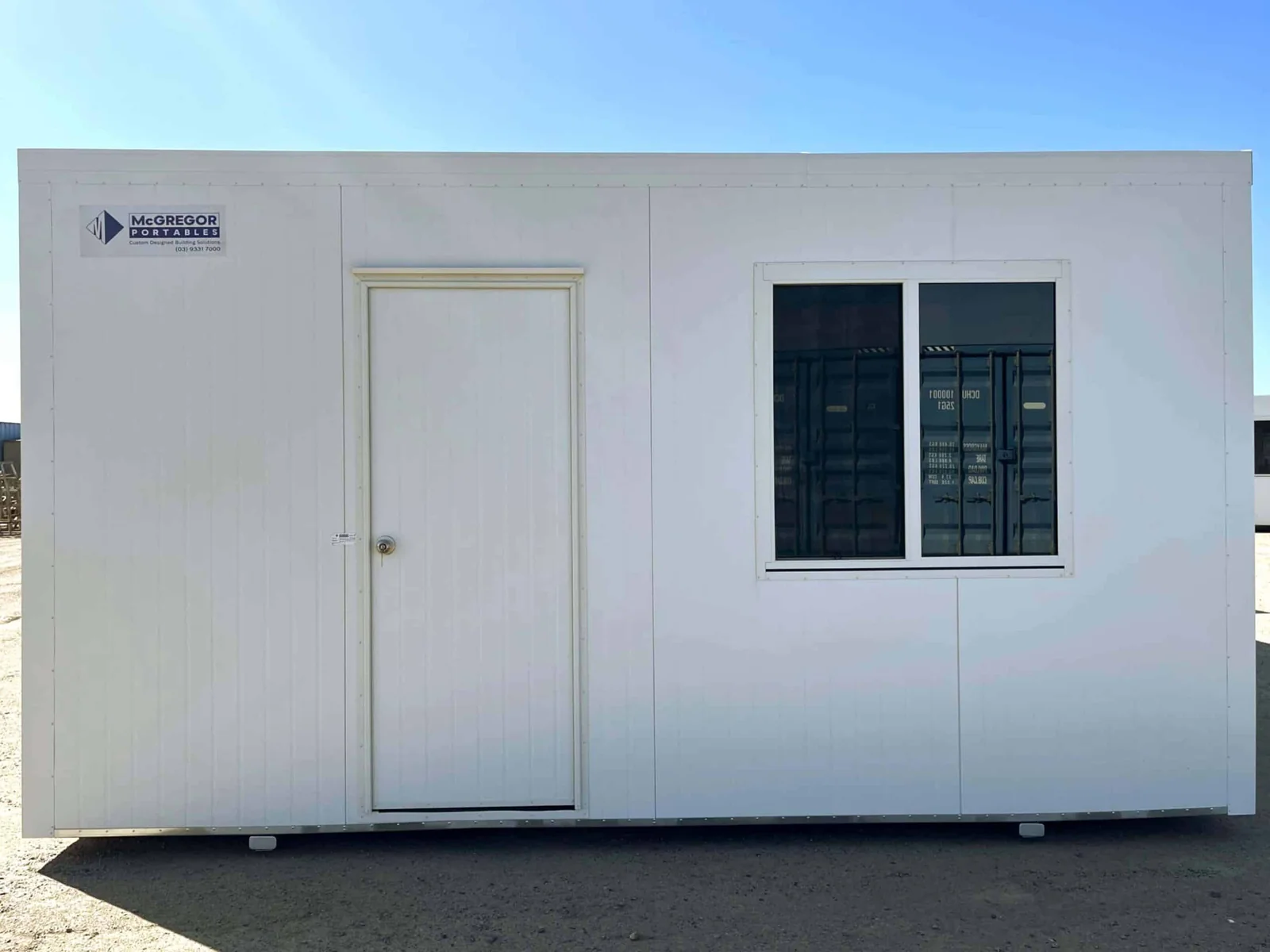 White site office with a single door and one security-screened window, on a gravel yard.
