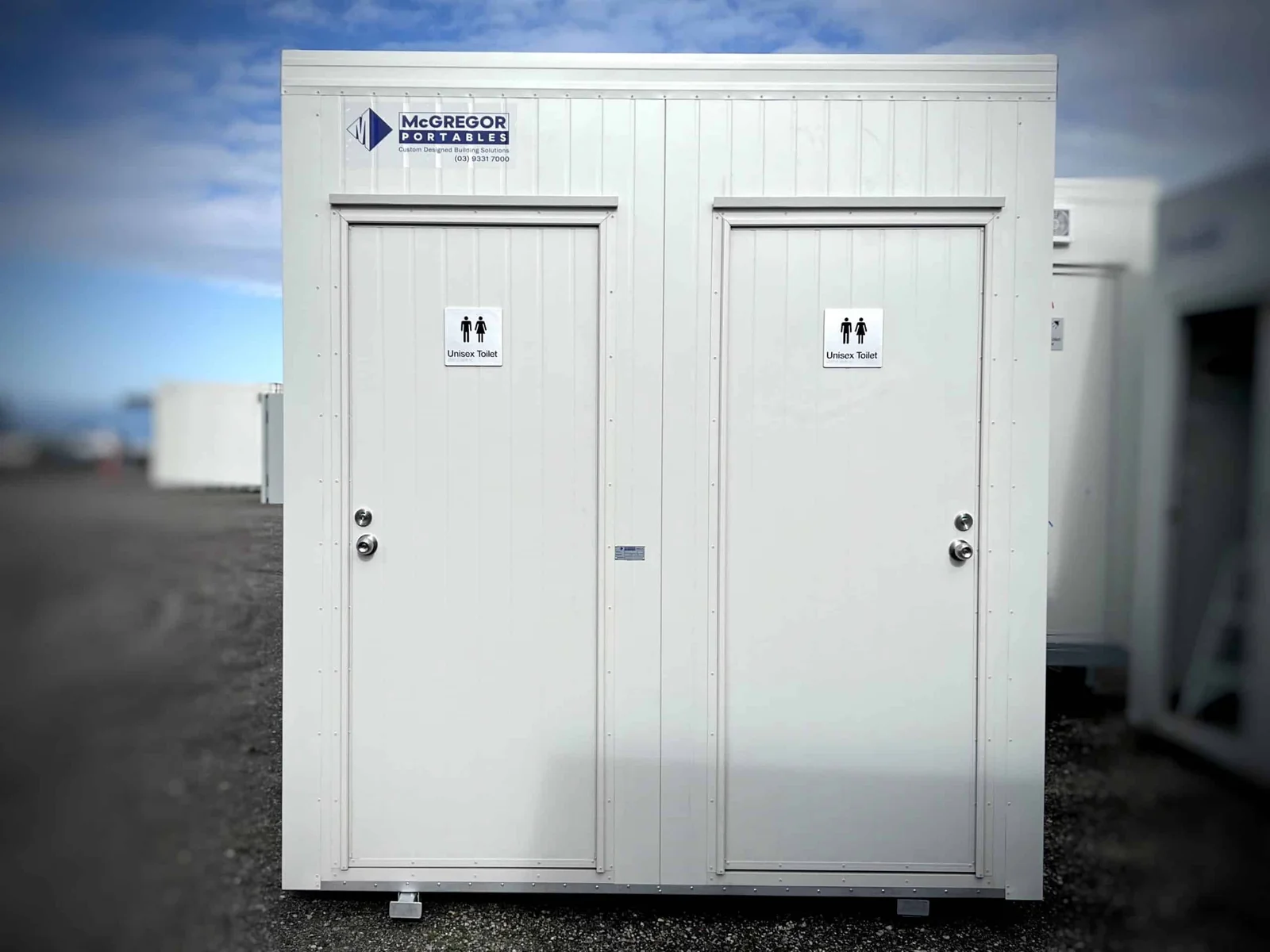 White twin toilet unit with two signed doors side by side, on gravel.