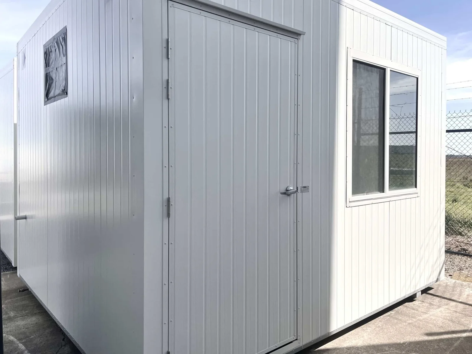 White first aid room with a single door and a security-screened window, beside a chain-link fence.