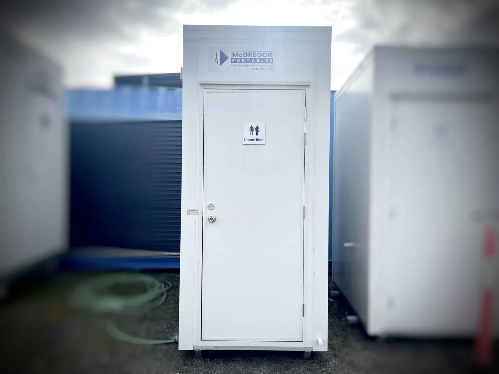 Single white toilet unit with one signed door, standing among other buildings in the yard.