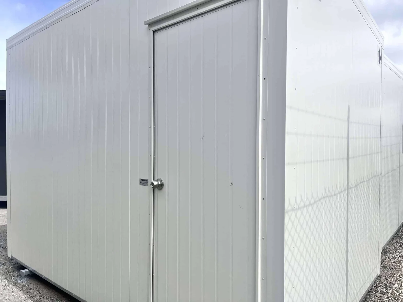 White site office with a single central door and blank cladding, beside a chain-link fence.