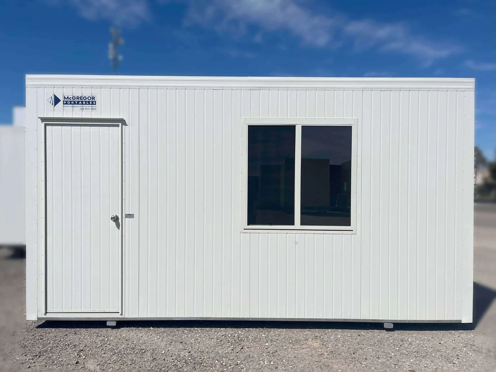 White site office with a single door and one large window, on a gravel yard under a blue sky.