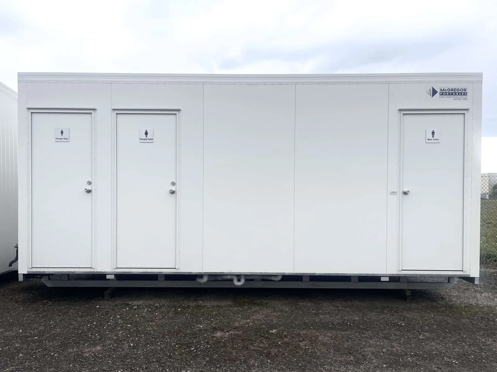 Long white toilet block with three signed doors along the front, mounted on a steel frame.