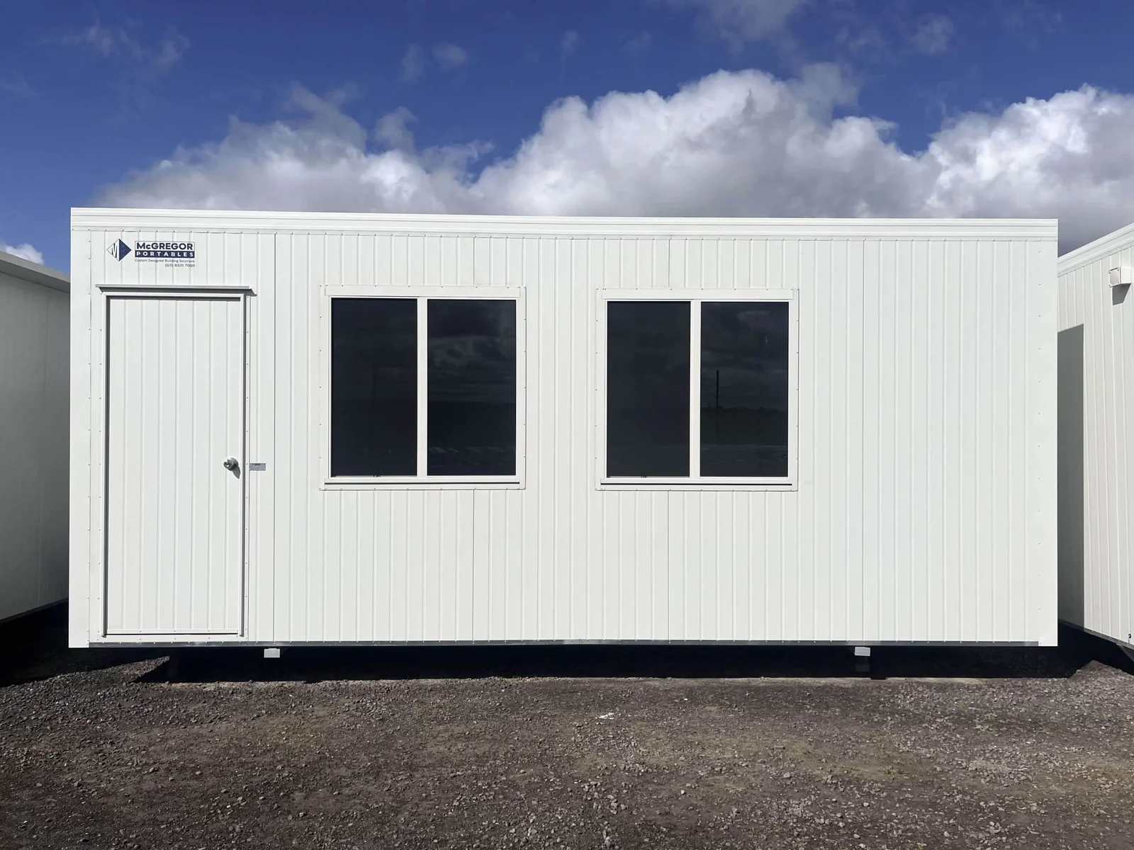 White site office with a door at one end and two large windows, on a gravel yard.