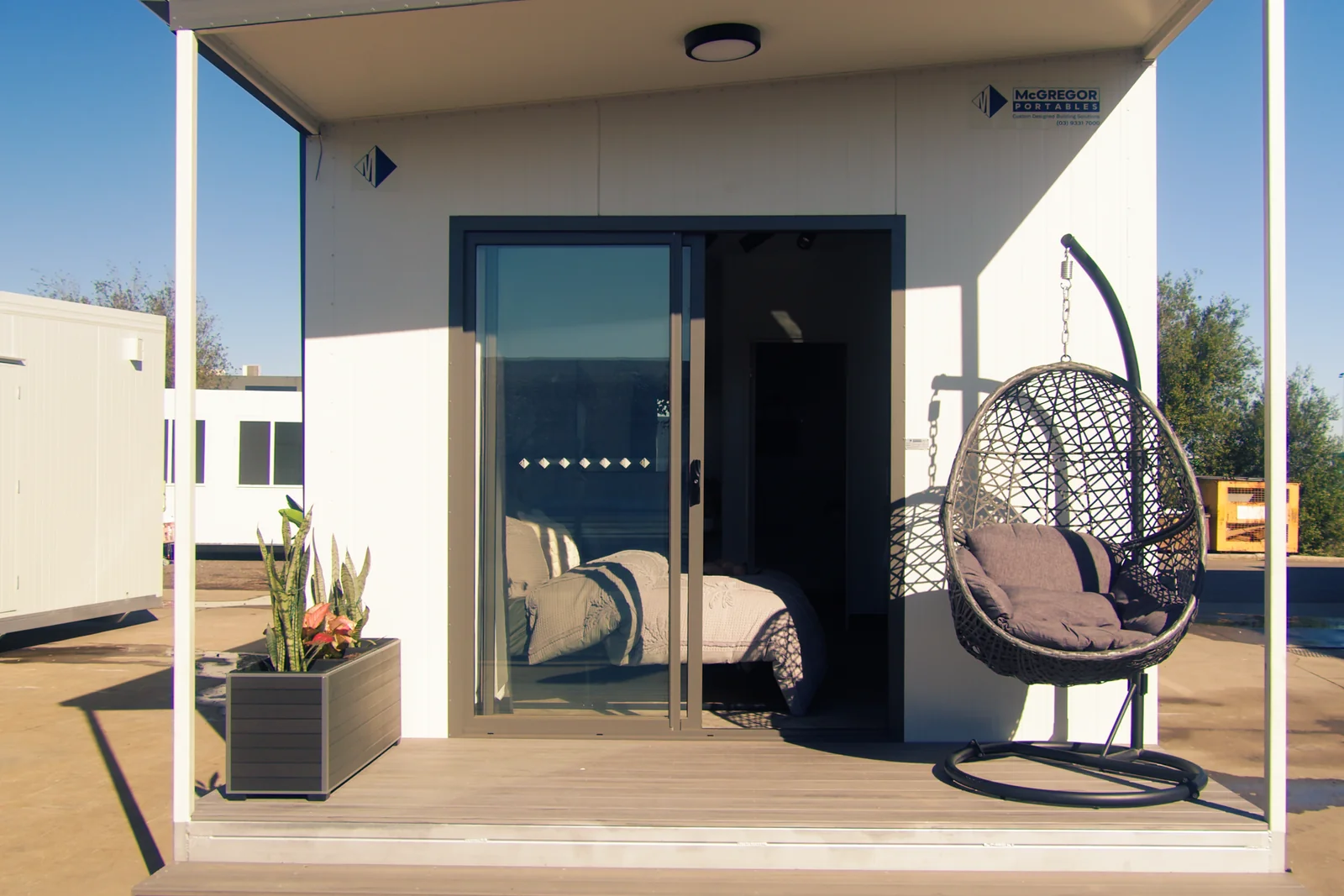 Studio accommodation unit with a sliding glass door, a timber deck, a hanging egg chair and planter boxes.