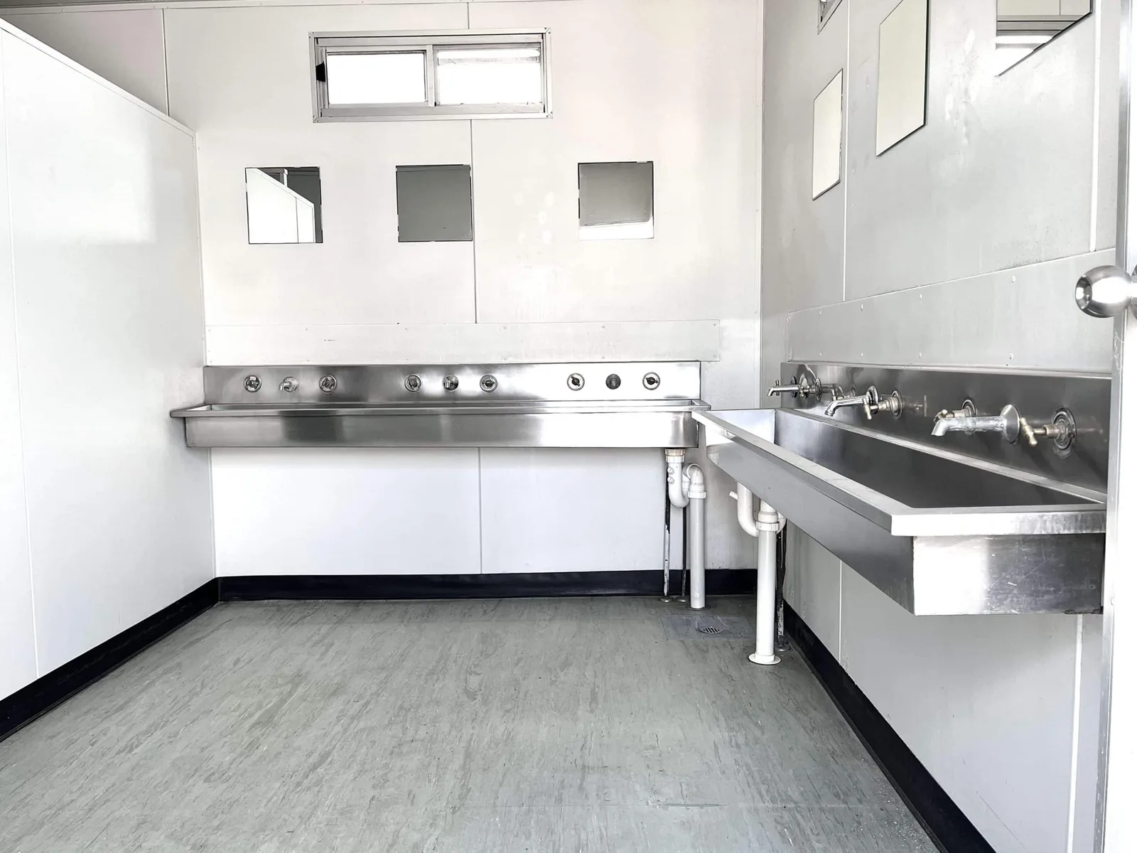 Interior of a portable ablution block with two stainless steel trough sinks, multiple taps and small mirrors on the wall
